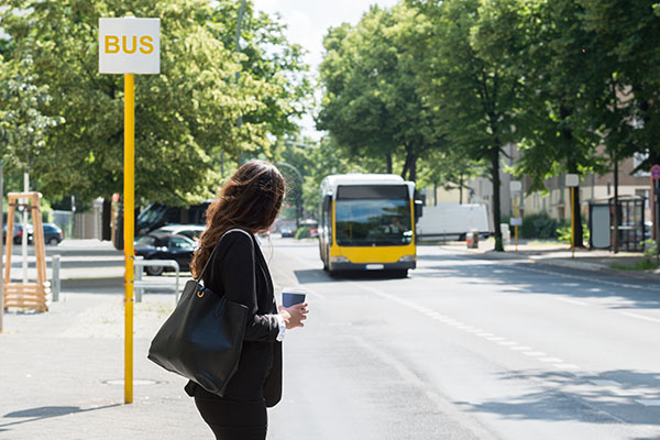 A woman waits at a bus stop holding a coffee cup, while a bus approaches on a tree-lined street.