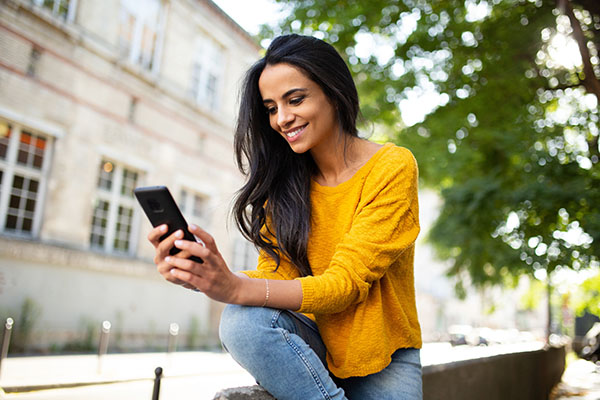 A woman wearing a yellow sweater and jeans sits outdoors, smiling while looking at her smartphone.