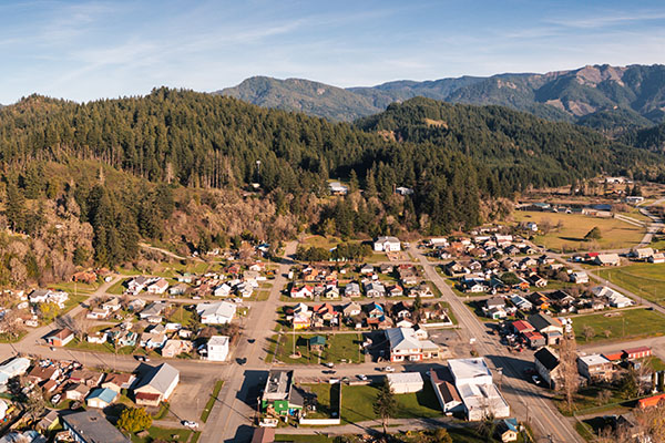 Aerial view of a small town with scattered houses, roads, and buildings, surrounded by forested hills and mountains under a clear sky.